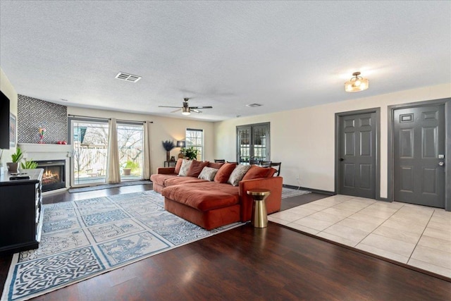 living area featuring a warm lit fireplace, a textured ceiling, wood finished floors, visible vents, and a ceiling fan