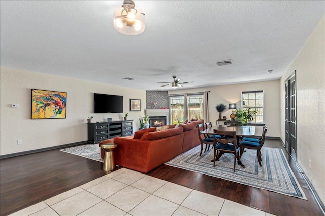 living area featuring a textured ceiling, wood finished floors, visible vents, a ceiling fan, and a lit fireplace