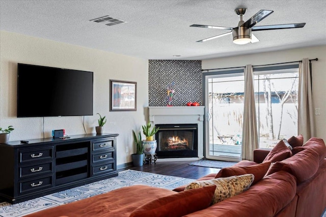 living area with a textured ceiling, wood finished floors, visible vents, a ceiling fan, and a glass covered fireplace