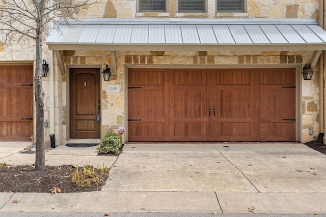 property entrance featuring a garage, stone siding, metal roof, and driveway