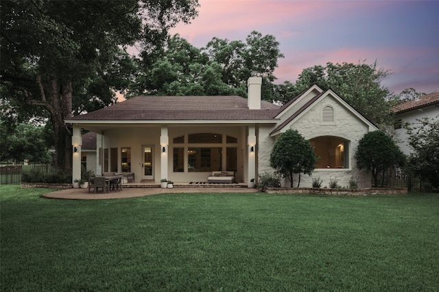 back of house at dusk featuring a patio area, a chimney, fence, and a yard