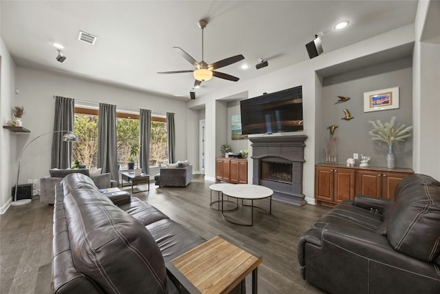 living area featuring a fireplace with raised hearth, a ceiling fan, visible vents, baseboards, and dark wood finished floors