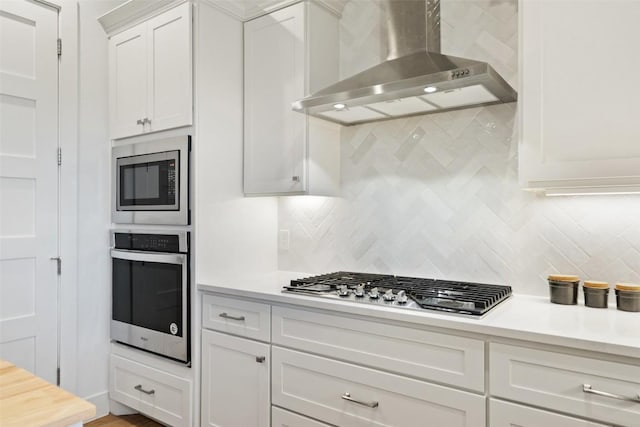 kitchen with tasteful backsplash, white cabinets, wall chimney exhaust hood, stainless steel appliances, and light countertops