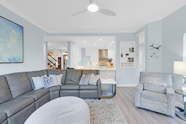 living room featuring ceiling fan, light wood-style flooring, baseboards, ornamental molding, and stairway