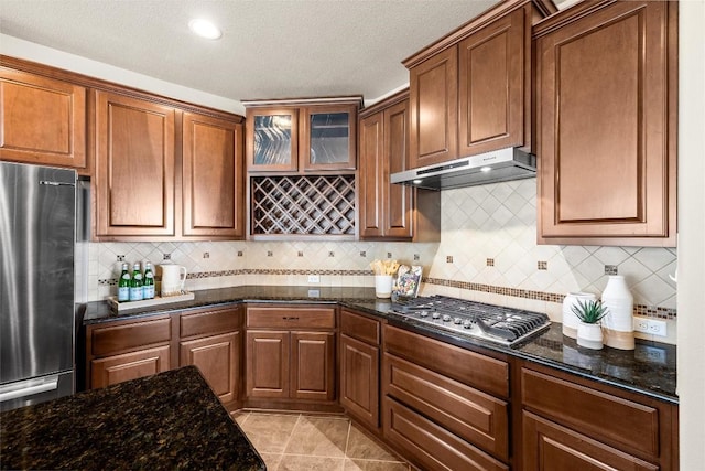 kitchen with brown cabinets, light tile patterned floors, stainless steel appliances, dark stone countertops, and under cabinet range hood