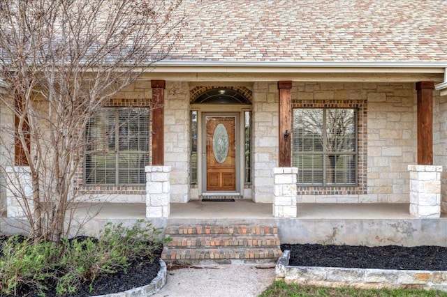 entrance to property featuring stone siding, a porch, and roof with shingles