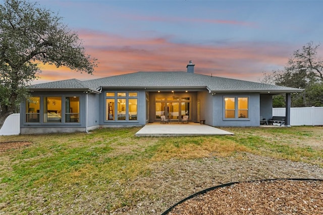 back of house at dusk with a patio, fence, a yard, stucco siding, and a chimney