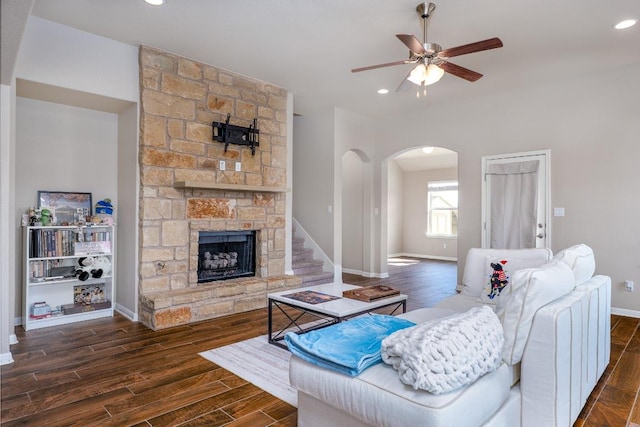 living room with arched walkways, dark wood-style flooring, a ceiling fan, a stone fireplace, and baseboards