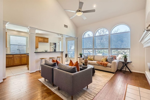 living area with high vaulted ceiling, light wood-style flooring, visible vents, a ceiling fan, and decorative columns