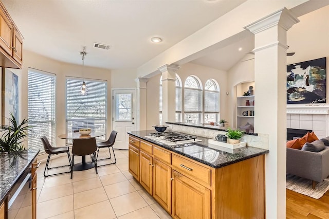 kitchen with built in shelves, gas cooktop, dark stone counters, a tiled fireplace, and decorative columns