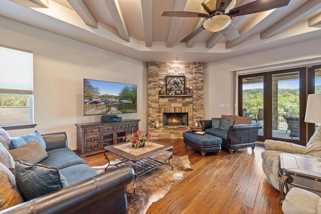 living area with ceiling fan, a stone fireplace, wood finished floors, and beam ceiling