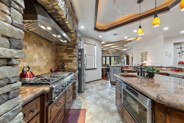 kitchen with stainless steel appliances, a sink, light stone countertops, wall chimney exhaust hood, and a raised ceiling