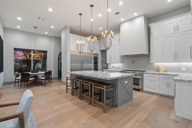 kitchen featuring custom exhaust hood, light wood-style flooring, a kitchen island with sink, high quality appliances, and tasteful backsplash