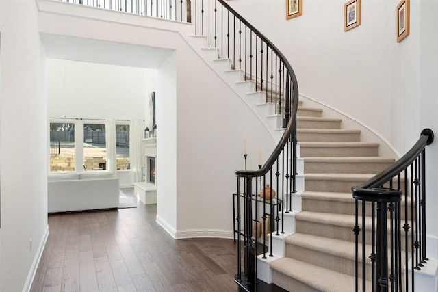 staircase with a warm lit fireplace, a towering ceiling, baseboards, and wood finished floors