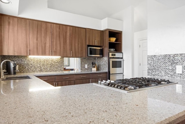 kitchen featuring tasteful backsplash, light stone counters, stainless steel appliances, open shelves, and a sink