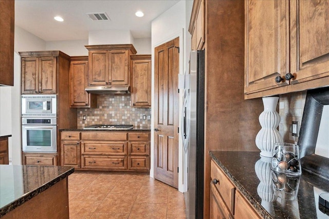 kitchen with visible vents, appliances with stainless steel finishes, brown cabinetry, dark stone countertops, and under cabinet range hood