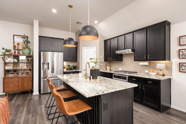 kitchen with under cabinet range hood, appliances with stainless steel finishes, light stone countertops, tasteful backsplash, and a kitchen bar