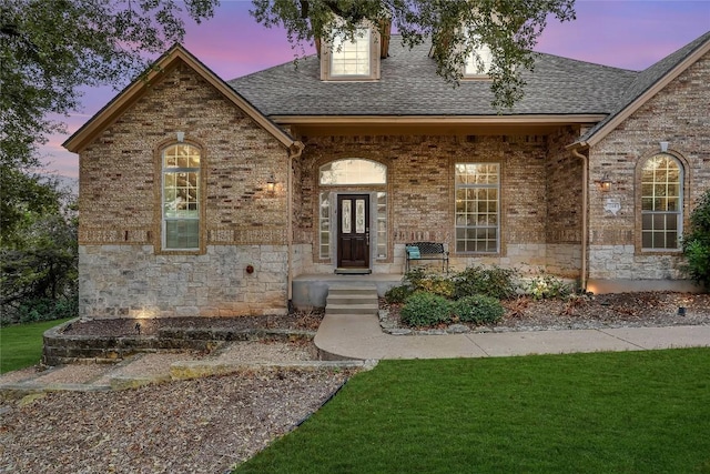 french provincial home with stone siding, a yard, roof with shingles, and brick siding