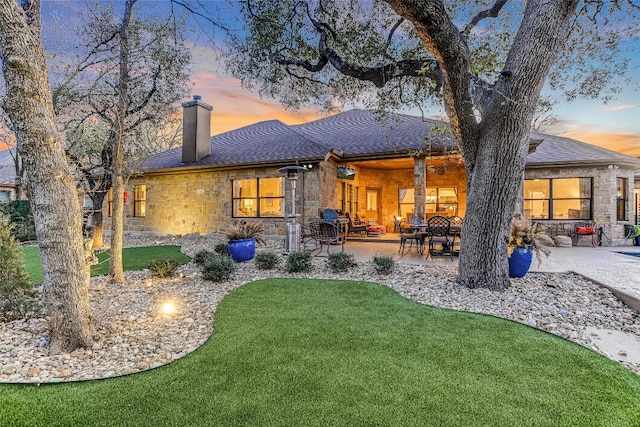 back of house at dusk featuring a lawn, stone siding, a chimney, roof with shingles, and a patio area