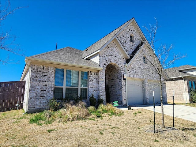 french provincial home with brick siding, concrete driveway, roof with shingles, and fence