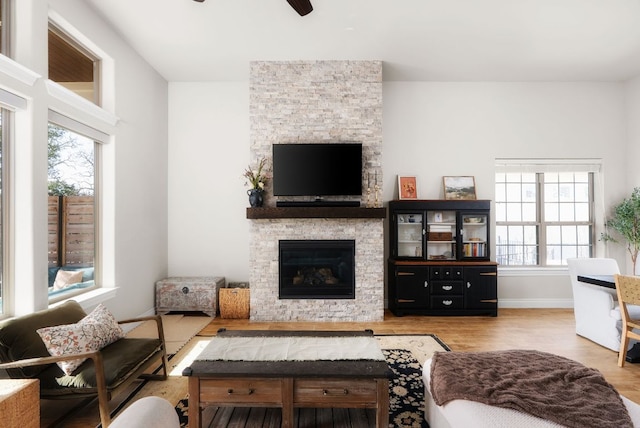 living area featuring a ceiling fan, a stone fireplace, baseboards, and wood finished floors