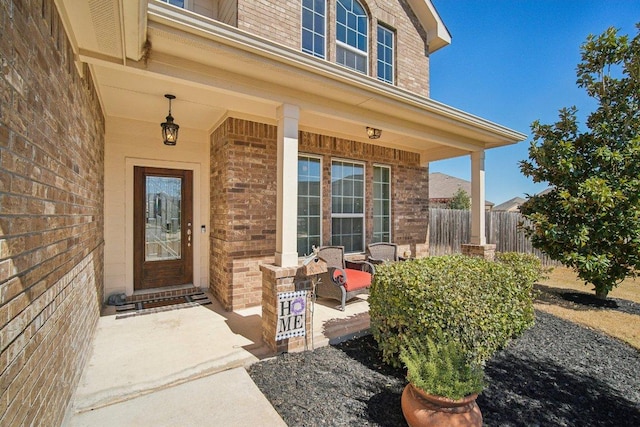 view of exterior entry featuring covered porch, brick siding, and fence