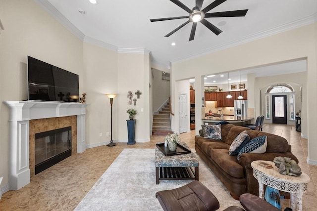 living area with stairway, ornamental molding, a ceiling fan, a tile fireplace, and baseboards