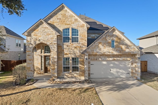 french country inspired facade with a garage, concrete driveway, stone siding, fence, and roof mounted solar panels