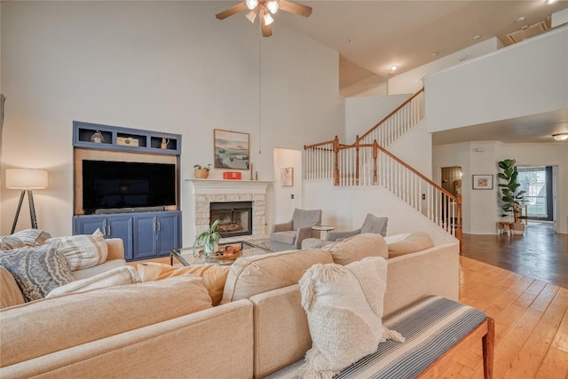 living area with a towering ceiling, hardwood / wood-style flooring, ceiling fan, stairs, and a stone fireplace