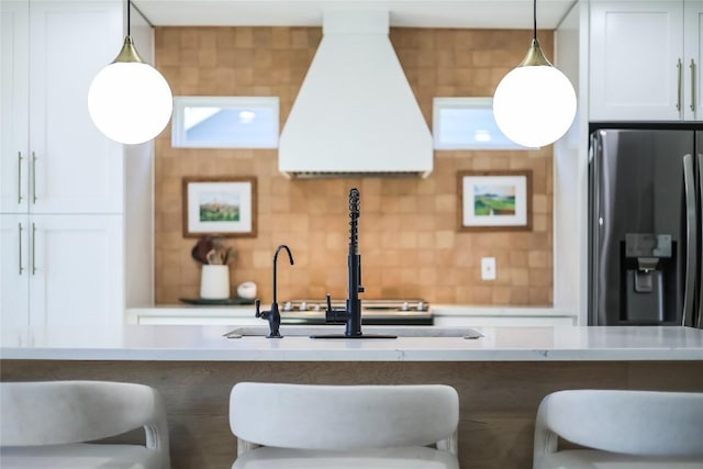 kitchen with stainless steel refrigerator with ice dispenser, a breakfast bar area, light countertops, decorative backsplash, and premium range hood