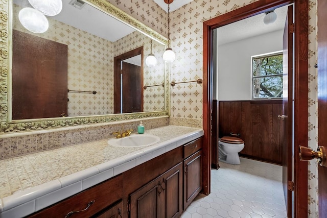 bathroom featuring wallpapered walls, wainscoting, toilet, tile patterned floors, and vanity