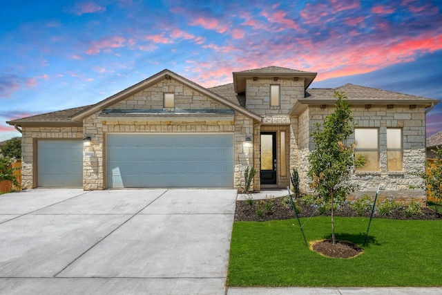 french country style house with roof with shingles, a yard, a garage, stone siding, and driveway