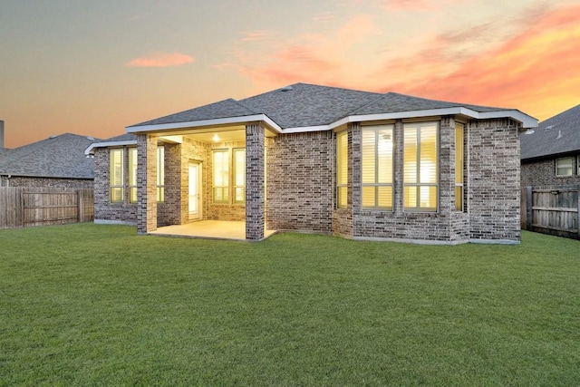 back of property at dusk featuring roof with shingles, a yard, brick siding, a patio, and a fenced backyard