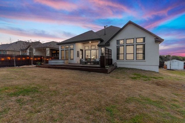 back of property at dusk with a deck, a jacuzzi, fence, a yard, and a shingled roof