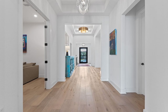 corridor featuring a tray ceiling, baseboards, light wood-style floors, and a notable chandelier