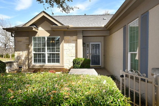 view of exterior entry with stone siding, fence, stucco siding, and roof with shingles