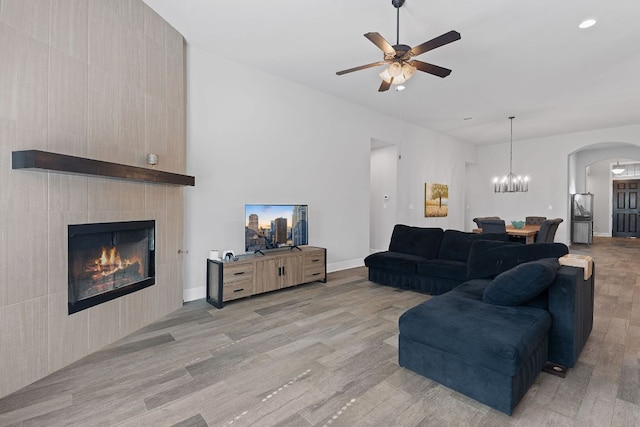 living area featuring arched walkways, ceiling fan with notable chandelier, baseboards, light wood-type flooring, and a tiled fireplace