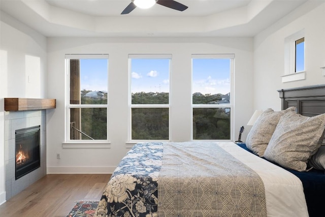 bedroom with a tiled fireplace, multiple windows, a raised ceiling, and wood finished floors