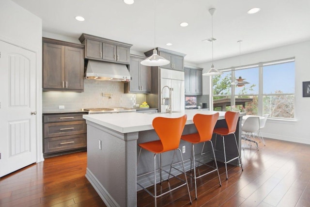 kitchen with dark wood finished floors, light countertops, backsplash, an island with sink, and exhaust hood