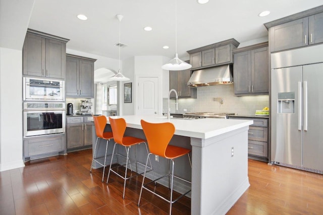 kitchen with built in fridge, a kitchen island with sink, under cabinet range hood, a sink, and tasteful backsplash