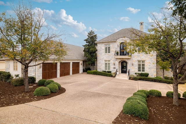 french country style house with a chimney, concrete driveway, a balcony, a garage, and stone siding