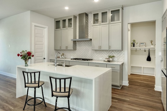 kitchen with decorative backsplash, baseboards, an island with sink, wall chimney exhaust hood, and a sink