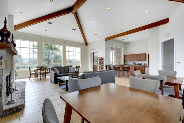 dining area with light tile patterned floors, visible vents, a stone fireplace, high vaulted ceiling, and beamed ceiling