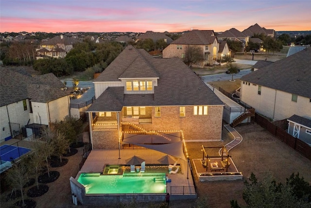 back of house at dusk with a shingled roof, a patio area, a residential view, and a hot tub