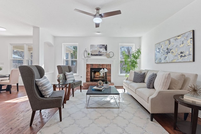 living room with ceiling fan, a brick fireplace, wood finished floors, and a healthy amount of sunlight