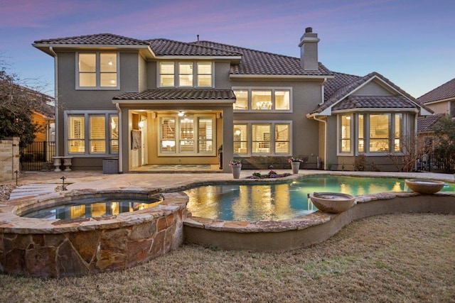 back of house at dusk with a patio, a pool with connected hot tub, stucco siding, a chimney, and a tile roof