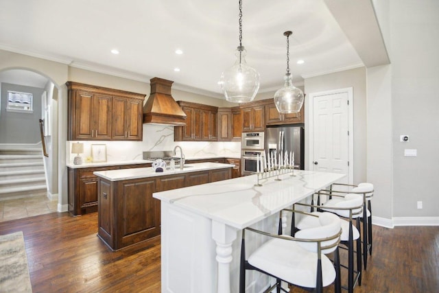 kitchen with dark wood-style floors, custom exhaust hood, appliances with stainless steel finishes, and an island with sink