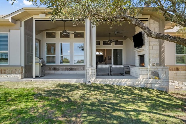 view of exterior entry with an outdoor stone fireplace, a yard, a patio, a ceiling fan, and stone siding