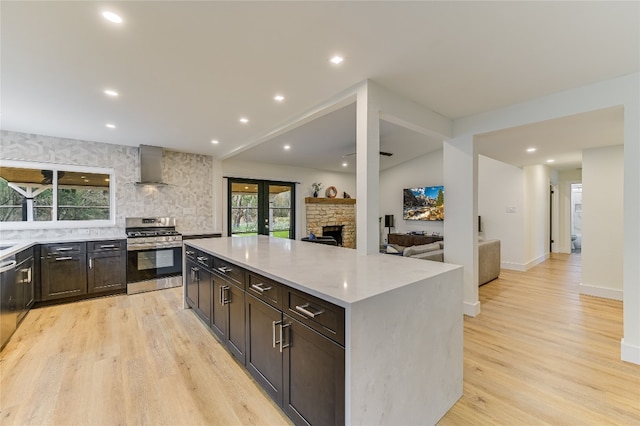 kitchen with stainless steel range with gas stovetop, a stone fireplace, wall chimney exhaust hood, and light wood-style floors