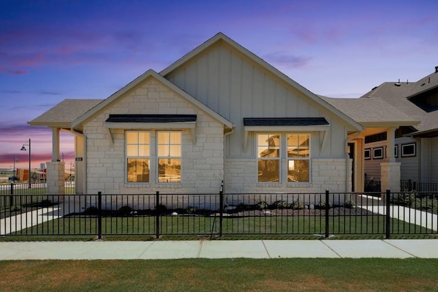 view of front of property featuring a shingled roof, stone siding, a fenced front yard, a yard, and board and batten siding